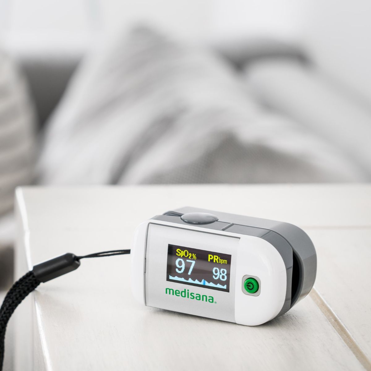 Ein weißes medisana Pulsoximeter liegt auf einem Tisch und zeigt auf dem Display die Messwerte für Sauerstoffsättigung und Puls an.